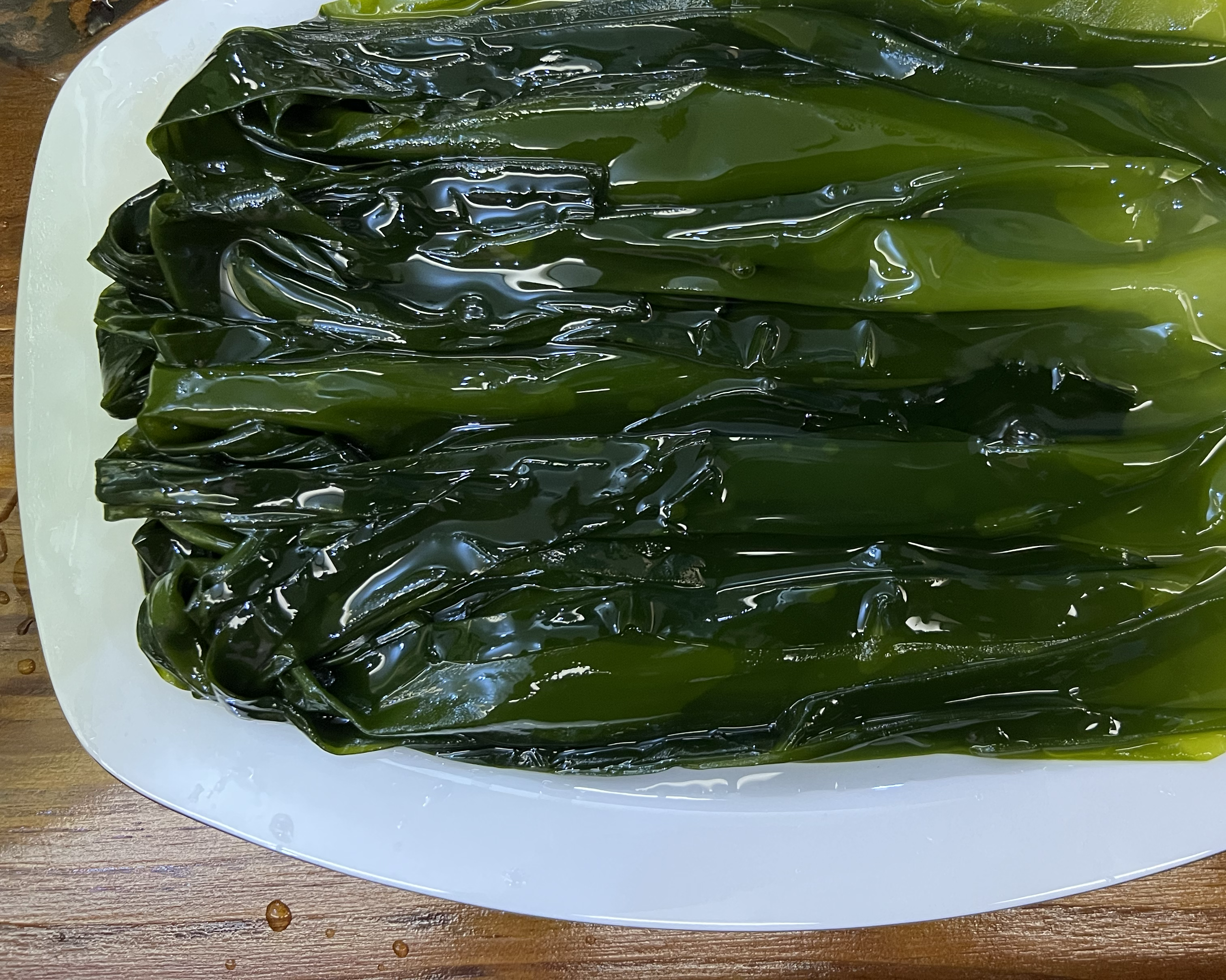 新鲜泡发后的深褐色海带叶片,表面光滑带有黏液,呈波浪状边缘,放置在白色瓷盘中,背景为厨房操作台环境 海带特写