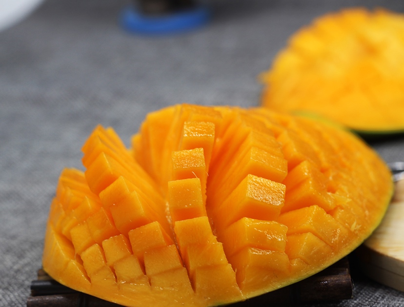 一个被横向切开的大青芒,露出金黄色的果肉,汁水丰盈,果核扁平光滑,背景为木质餐桌和绿叶衬托,突出果实的新鲜与诱人质感 切开的大青芒特写展示果肉与果汁