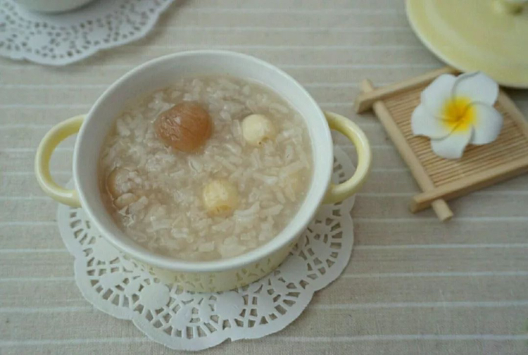 生活场景图:70岁左右中国城市老年女性双手捧青花瓷碗,专注食用温热的湘莲银耳羹;碗内莲子粒粒分明、软而不烂,银耳胶质丰盈;背景桌面可见湘莲真空包装袋(印有‘国家地理标志’‘湘潭湘莲’字样)及电子血压计屏幕显示‘128/76 mmHg’,强化真实健康场景 老年人手捧瓷碗食用莲子银耳羹特写:碗中为炖煮软糯的湘莲与银耳,莲子呈半透明乳白色,银耳舒展如云朵,汤汁清亮微稠,老人手腕佩戴智能血压计,桌面散落莲子干品与营养标签