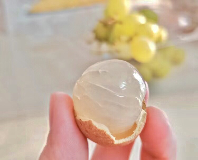 近距离拍摄剥壳后的石硖龙眼果肉,呈乳白色半透明状,表面光滑有弹性,光线照射下略带珠光质感。背景虚化显示绿色叶片,突出天然生态种植环境 石硖龙眼果肉特写高清图