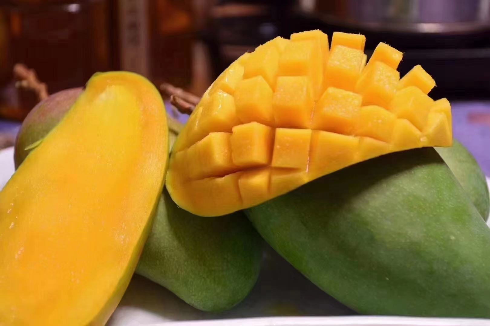 一张高清近距离拍摄的切开后的小台农芒,露出橙黄色的细腻果肉,汁水丰盈,刀口处可见光滑无丝的质地。旁边摆放完整未切开的果实,表皮呈亮金色略带淡红晕,背景为木质托盘与绿叶装饰,突出水果的新鲜与高品质感 小台农芒果实特写展示图