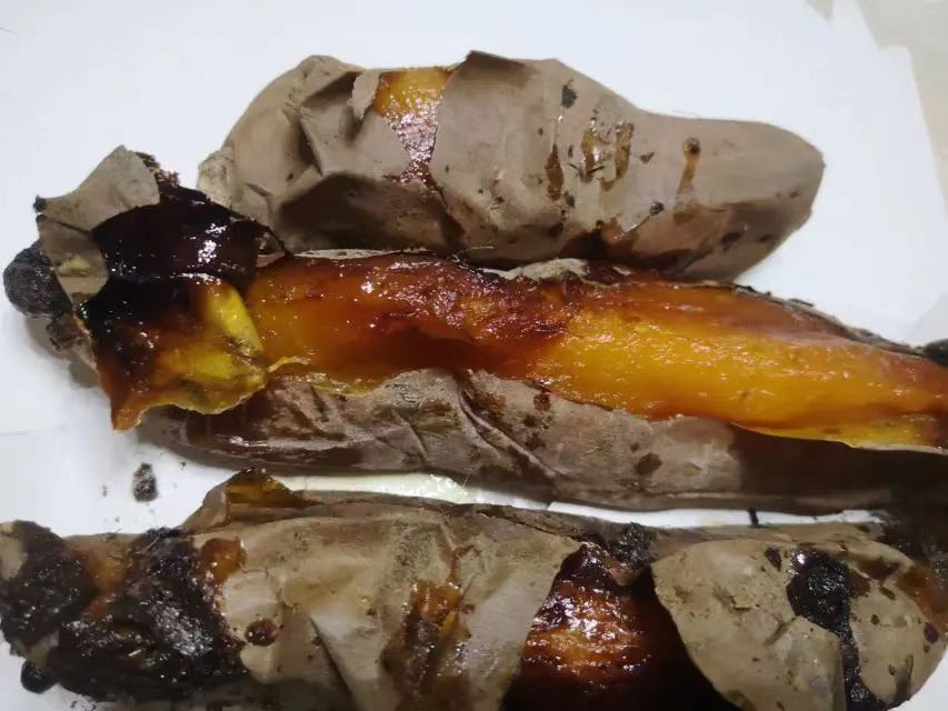 一个被横向切开的烤烟薯25号,内部果肉呈金黄色,湿润有光泽,表面可见明显糖油渗出,边缘微焦,散发热气,旁边配有勺子和纸巾,展示即食场景 烤制后的烟薯25号切开特写