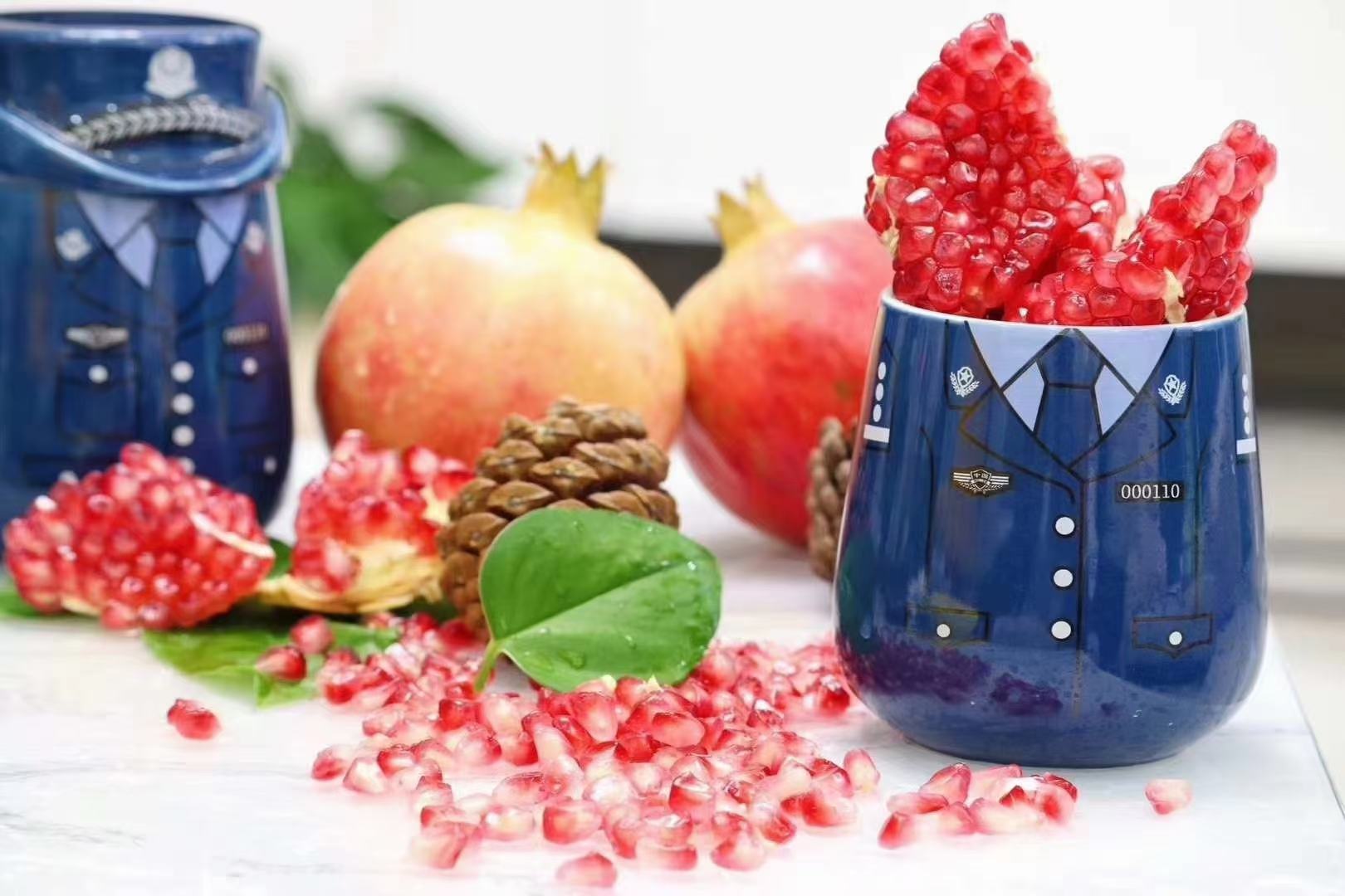 会理市石榴文化馆临展厅实景。中央U型木质展台陈列多组石榴主题文创:左侧为匠人现场雕刻的镂空石榴木雕;中部为民族银匠打造的石榴籽造型银耳坠与胸针;右侧为玻璃罐装‘石榴一’高端深加工产品线,含深红色石榴原浆、粉红色冻干脆片、琥珀色石榴籽油;背景墙为六尺宣纸绘制的工笔重彩《会理石榴四季图》,展现春花、夏荫、秋实、冬藏及采摘、分选、运输、品鉴场景 会理石榴文化主题展陈现场:木质展台上陈列手工石榴雕刻艺术品、石榴籽镶嵌银饰、水墨石榴书画长卷,以及透明罐装的‘石榴一’系列深加工产品(石榴原浆、冻干脆片、籽油礼盒),背景墙为巨幅手绘《会理石榴四季图》