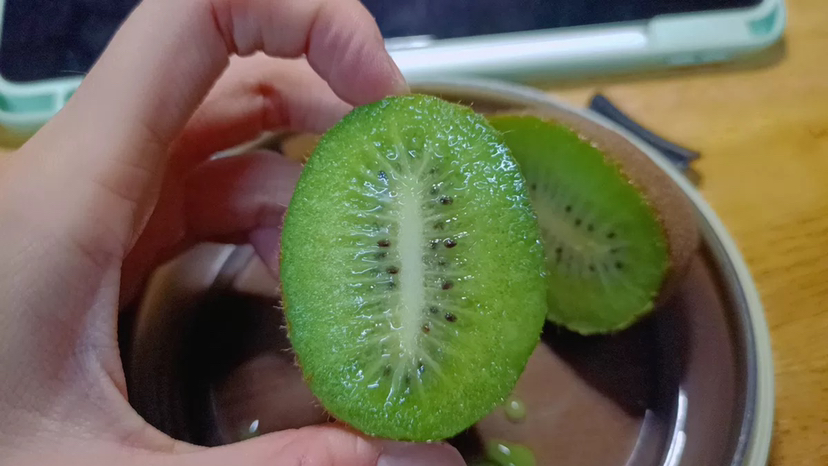 一颗成熟的徐香猕猴桃被横向切开,露出鲜艳的绿色果肉,周围排列着细密的黑色种子,果皮呈褐色绒毛状,背景为自然果园环境 徐香猕猴桃果实特写