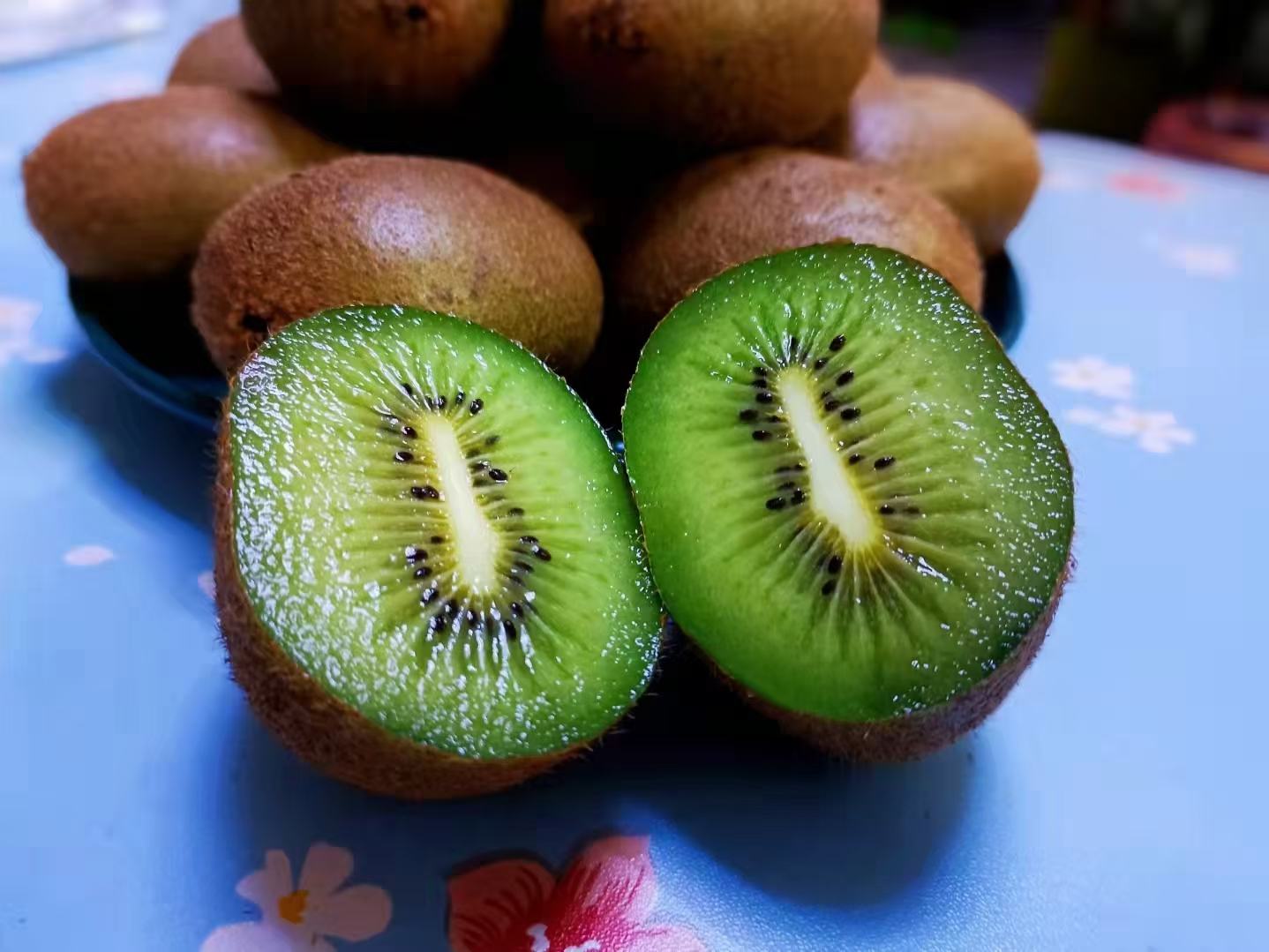 一颗被横向切开的翠香猕猴桃,露出鲜艳的翠绿色果肉,中心为放射状白色果心,果汁饱满,边缘带有细微水珠,旁边配有白色瓷盘和银色小勺,背景虚化显示木质餐桌与自然光线 切开的周至翠香猕猴桃特写