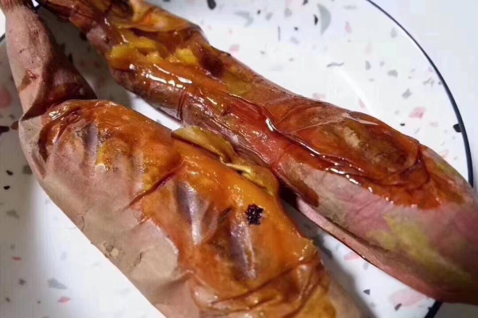 一只正在炭火炉中烘烤的烟薯25,外皮微焦裂开,露出金黄流蜜的内瓤,热气腾腾,周围摆放着几只已完成烤制、正在包装的烟薯,背景为街边小吃摊环境 烤制中的烟薯25特写