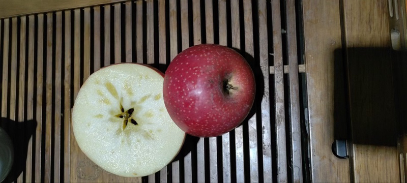 一张高清特写照片,展示三颗完整的花牛苹果并列摆放,果面呈现明显的五棱高桩形状和紫红色条纹,其中一颗被横向切开,露出乳白色果肉和饱满种子,背景为木质托盘和绿叶衬托 花牛苹果特写展示图