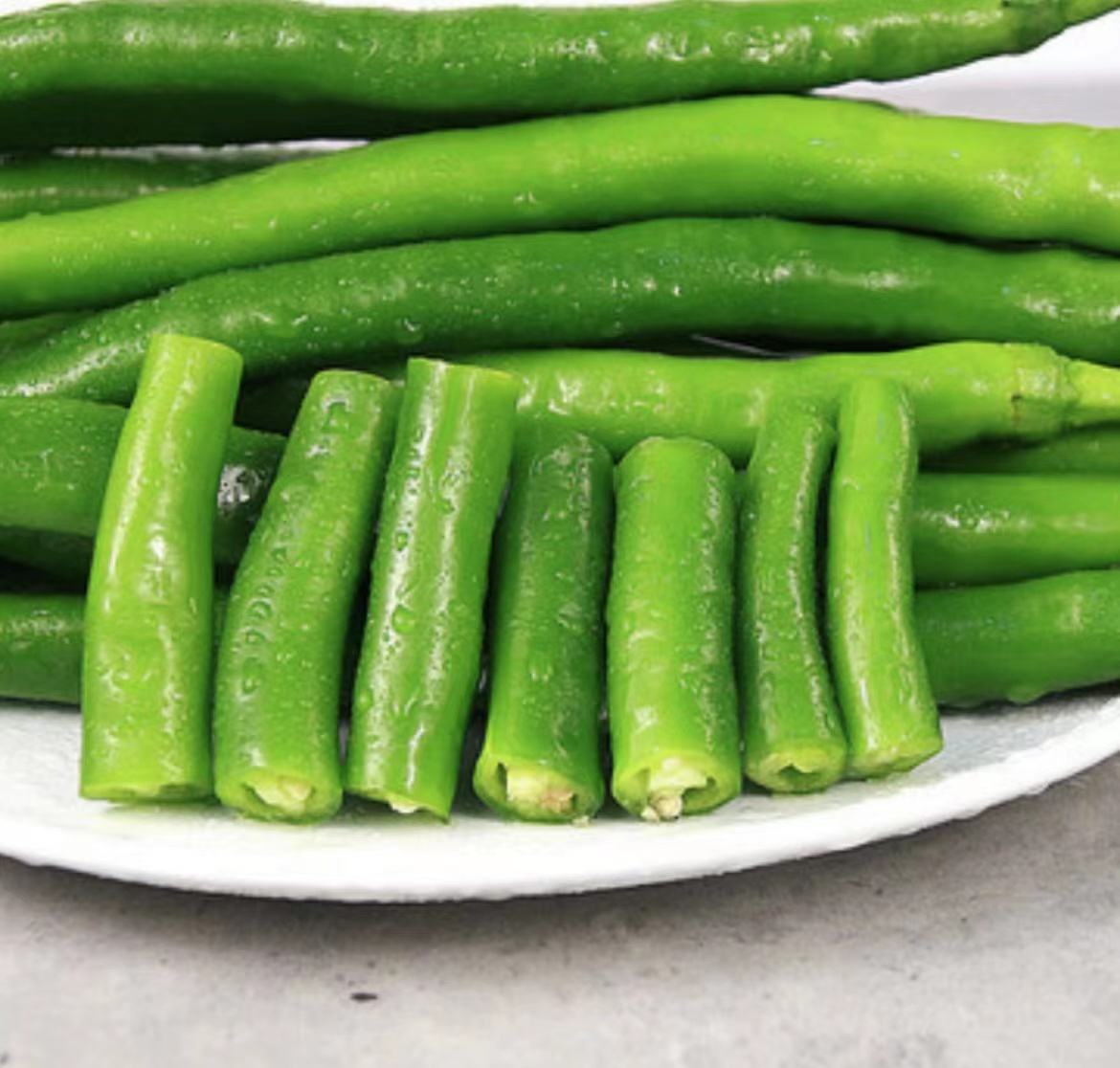 一张俯拍的厨房操作台照片,展示三种状态的青线椒:左边是完整的新鲜青线椒堆叠成小山状;中间是切成圈状的青线椒排列在白色瓷盘中;右边是经过油煸后的虎皮状青线椒,表面微焦起泡。背景有蒜瓣、姜片和酱油瓶,体现多种烹饪可能性 厨房中青线椒的不同处理方式