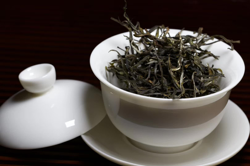 一个白瓷盖碗中正在冲泡的小青柑普洱茶,小青柑已开顶嵌入普洱茶团,热水注入后茶汤呈琥珀色,旁边配有茶夹、茶针和干茶样品,桌面铺有竹制茶席,营造出传统中式茶道氛围 小青柑与普洱茶组合冲泡过程