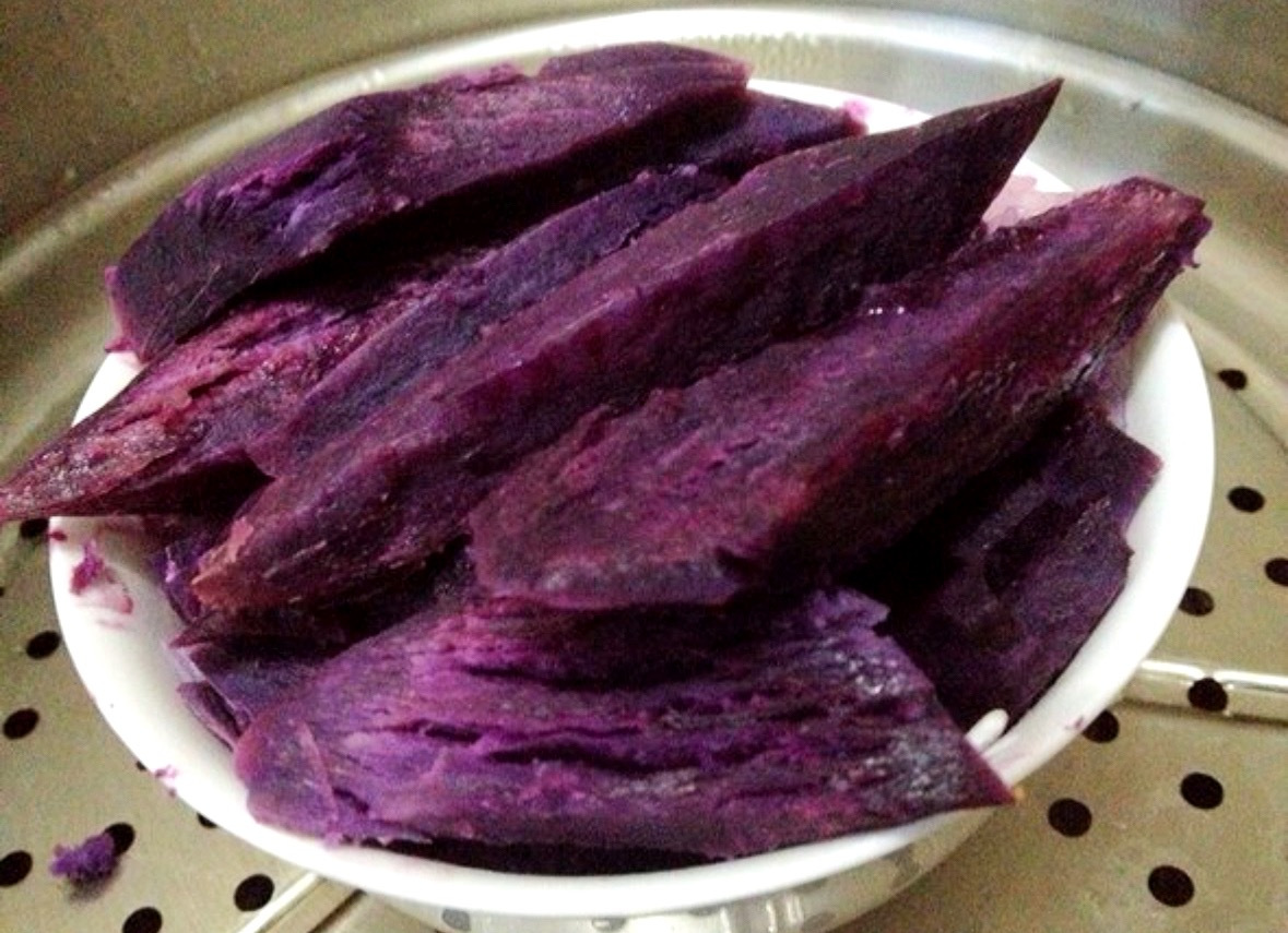 特写镜头：不锈钢勺舀起一勺红薯胡萝卜泥，泥体呈暖橙渐变色（红薯深橙过渡至胡萝卜亮橙），表面浮有芝麻粒与迷迭香碎，勺边凝结细小油珠，背景虚化处可见中国食物成分表（第6版）封面